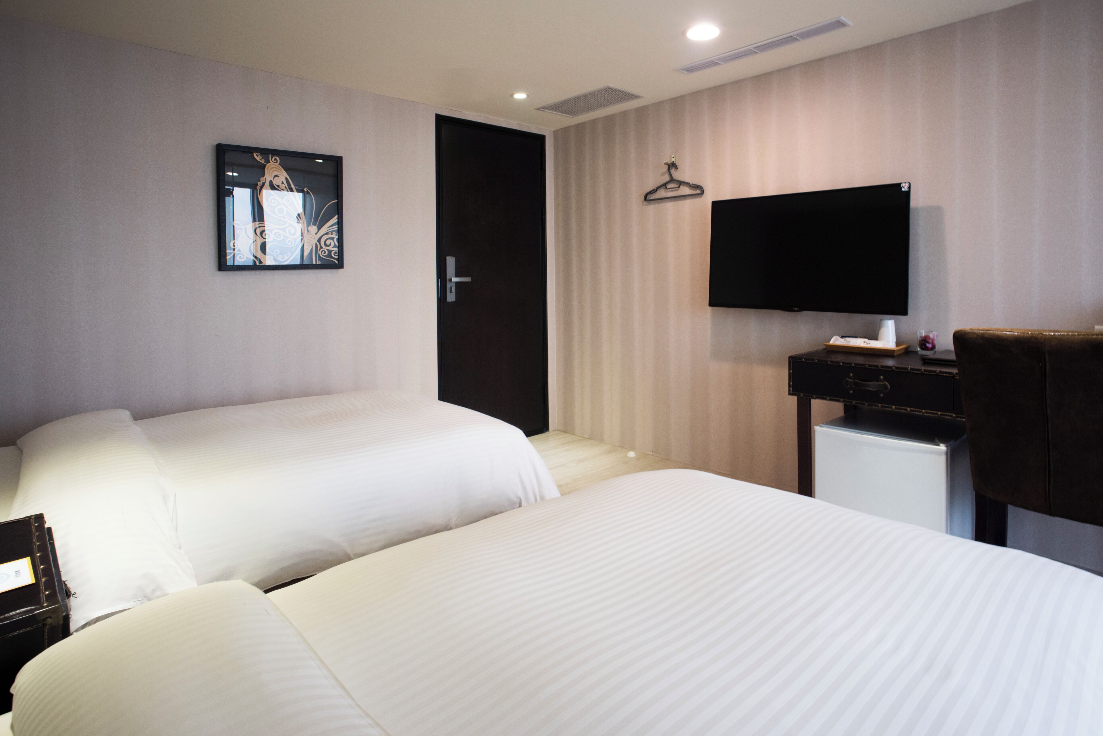 A hotel room with two beds, a flat-screen TV mounted on the wall, a small desk with a chair, and a door.