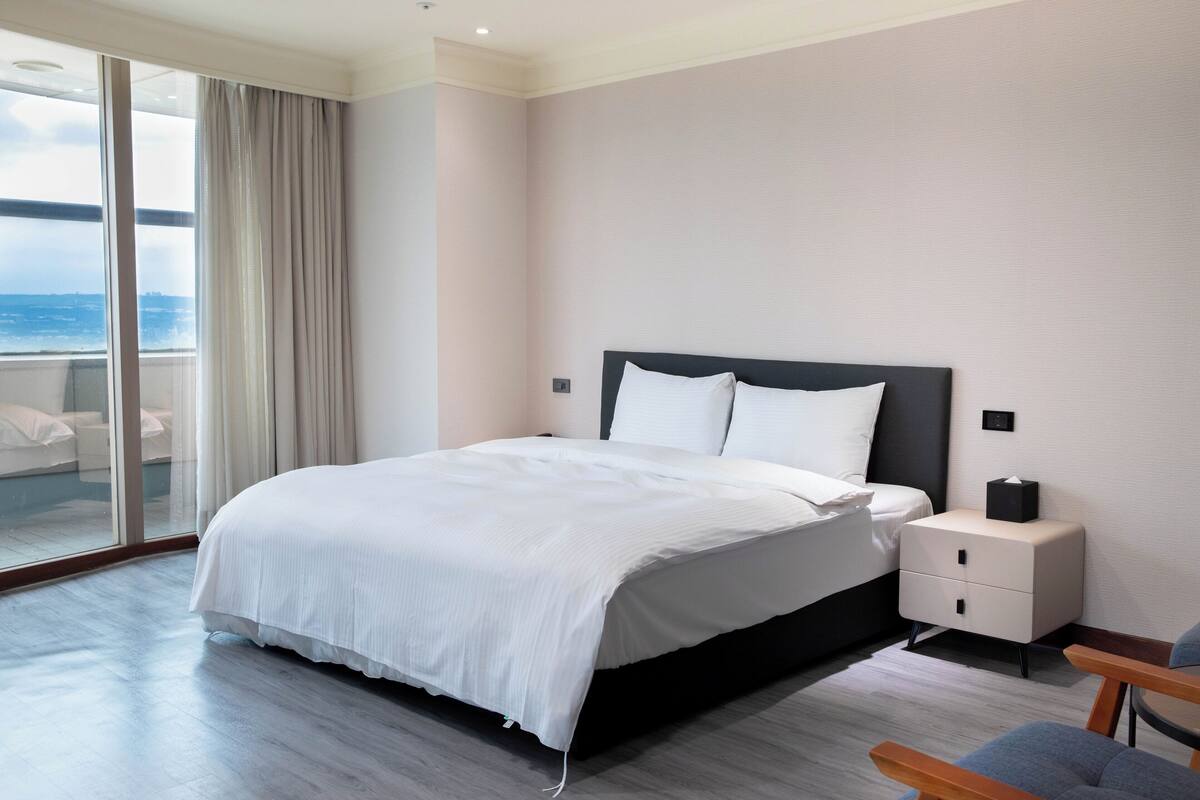 Une chambre d’hôtel moderne avec un grand lit, une table de chevet et une vue sur la mer, accessible par une porte-fenêtre coulissante.