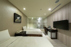 A hotel room with two beds, a desk, a chair, a flat-screen TV, and a glass shower enclosure.