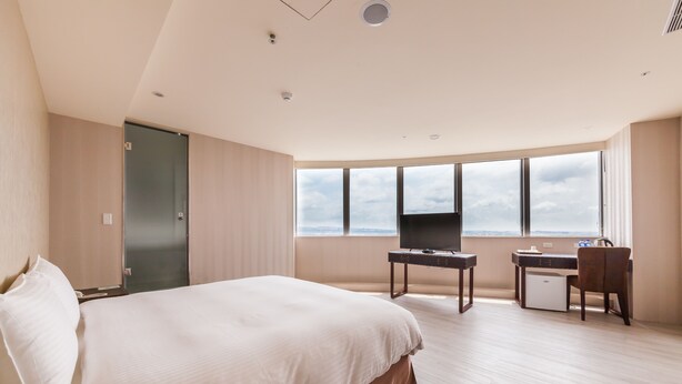 Een hotelkamer met een groot bed, een bureau met stoel, een televisie en uitzicht op de lucht door grote ramen.