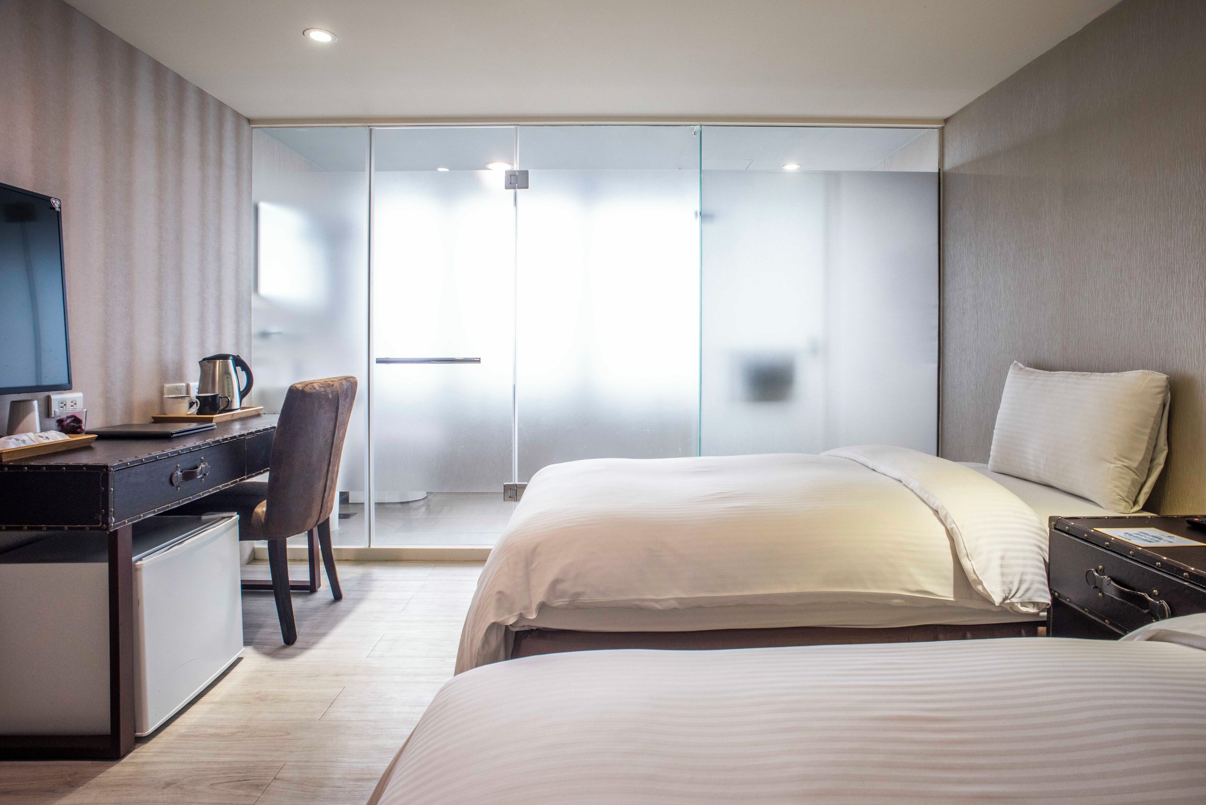 Habitación de hotel moderna con escritorio, silla, dos camas, televisor y puerta corrediza de vidrio al balcón.