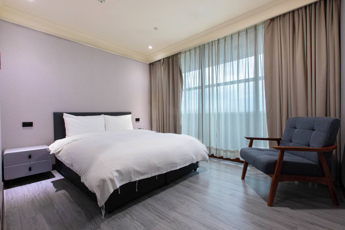 Une chambre à coucher avec un grand lit, une table de chevet en bois, une chaise et une fenêtre avec des rideaux.