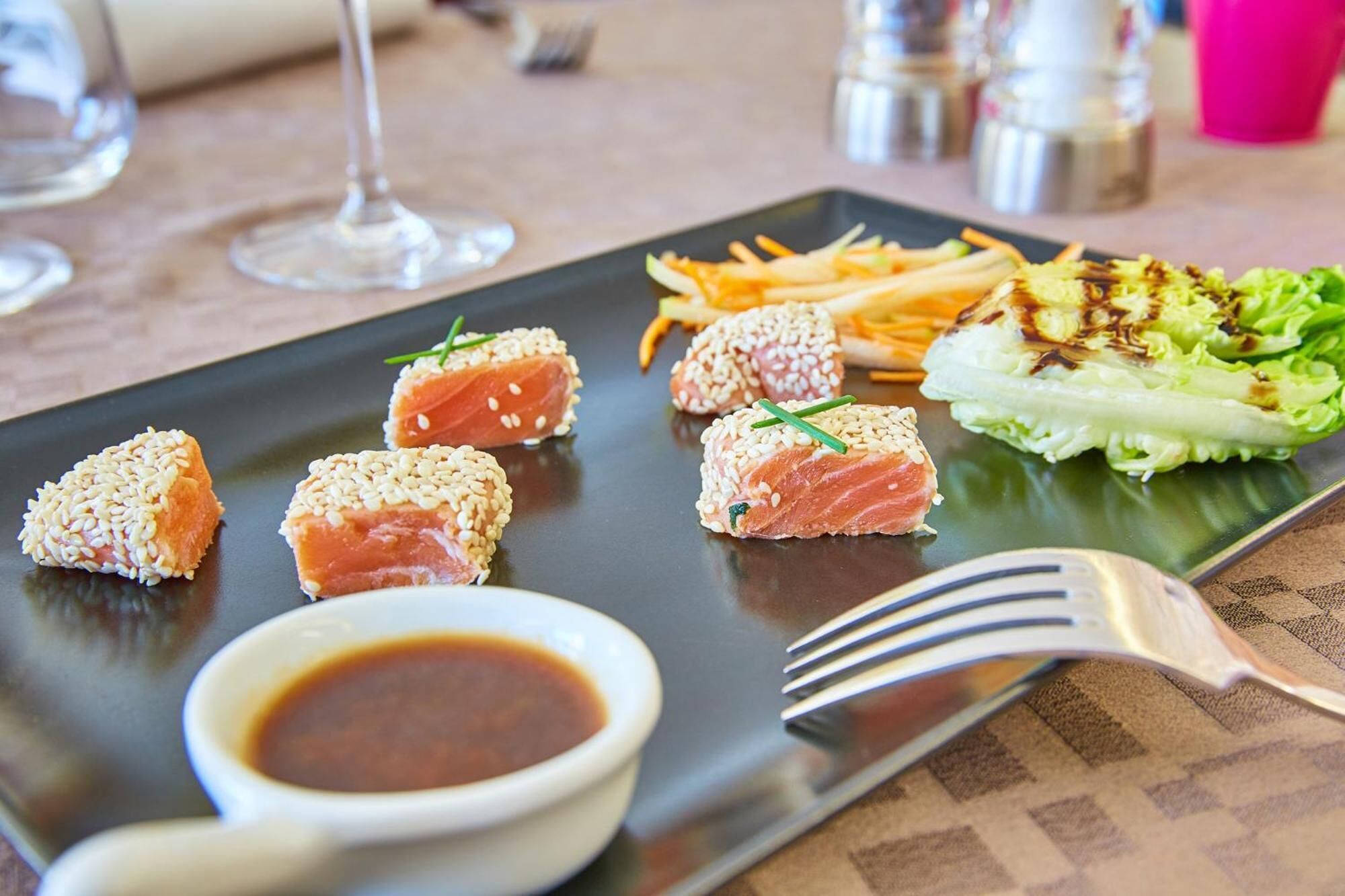 A black plate with sushi, a small bowl of sauce, and a fork on a table with a napkin.