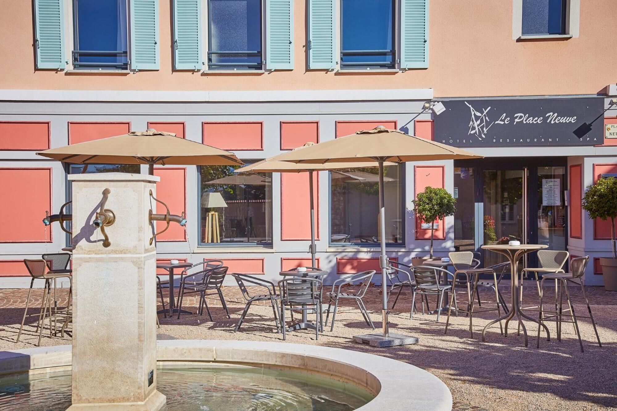 An outdoor seating area with tables and chairs, a fountain, and a building with a sign that reads "Le Place Nouvelle HOTEL RESTAURANT SPA".