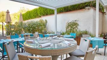 Un espace extérieur aménagé en terrasse, avec des tables rondes en verre et des chaises en osier, entouré d’un mur blanc et de verdure.