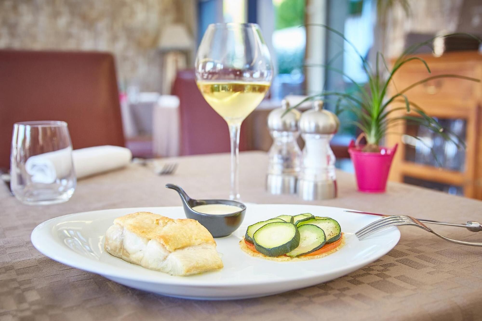 A plate with fish, a slice of cucumber, and a glass of white wine on a table.