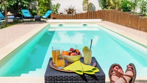 Un espace aménagé au bord de la piscine, avec une table sur laquelle sont disposées des boissons et des fruits, une chaise en osier et une vue sur la piscine et le jardin.