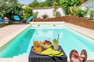 Um ambiente à beira da piscina com uma mesa com bebidas e frutas, uma cadeira de vime e vista para a piscina e o jardim.