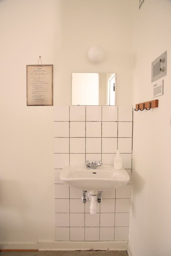 A bathroom with a white sink, a mirror above it, and a tiled wall. - SleepIn FÆNGSLET - Hostel (Horsens)