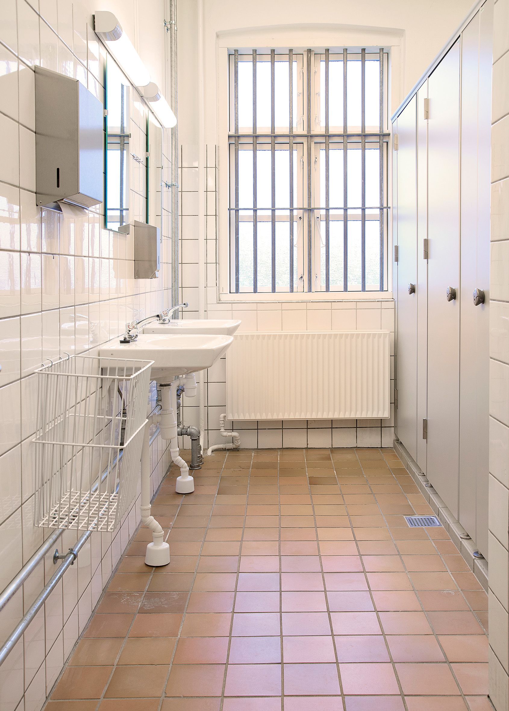Une salle de bain avec des murs recouverts de carreaux blancs, une fenêtre avec des barreaux, un lavabo, un miroir et un radiateur.