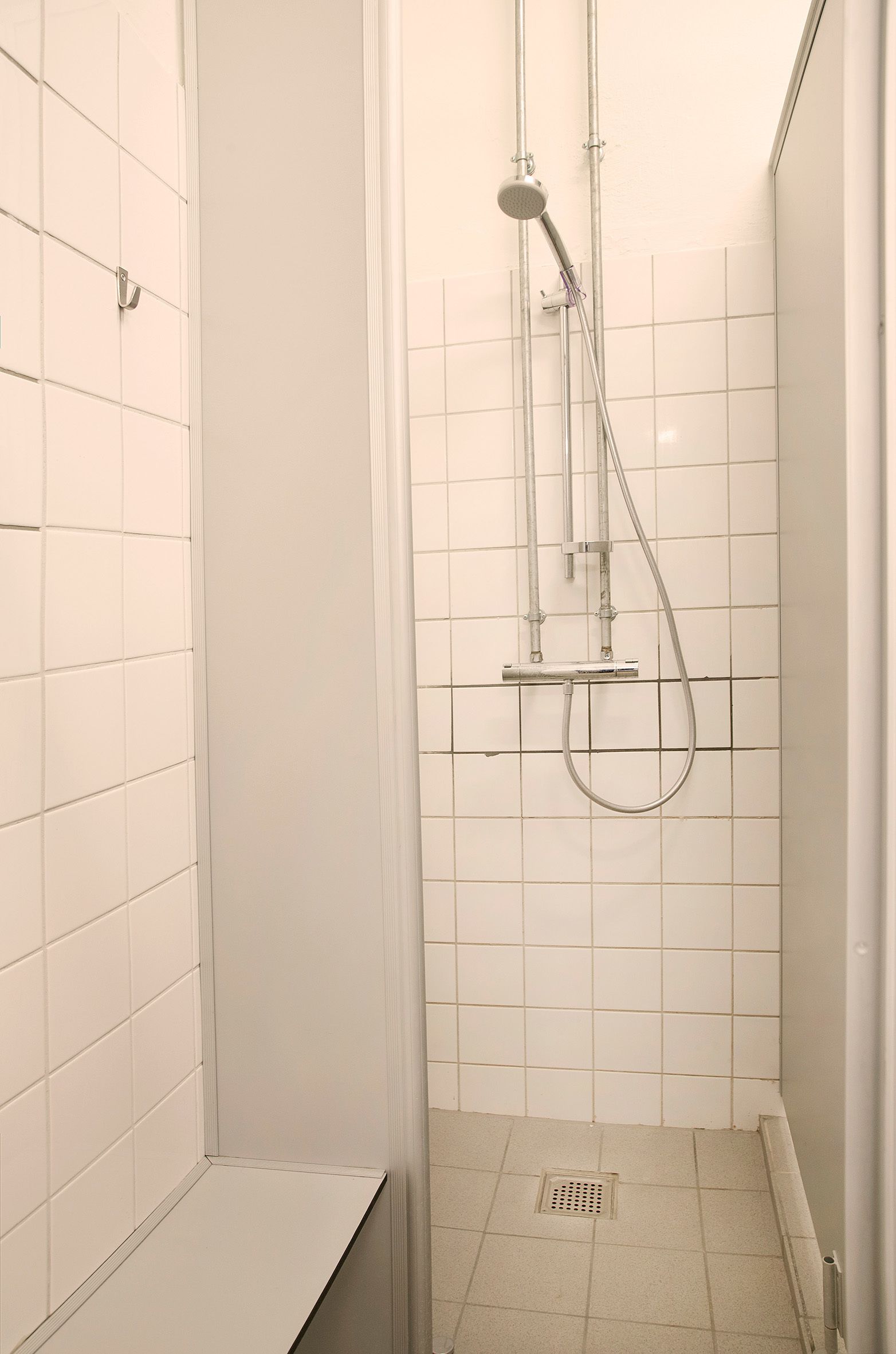 A small, enclosed shower area with white tiled walls and floor, a showerhead, and a handheld shower attachment.