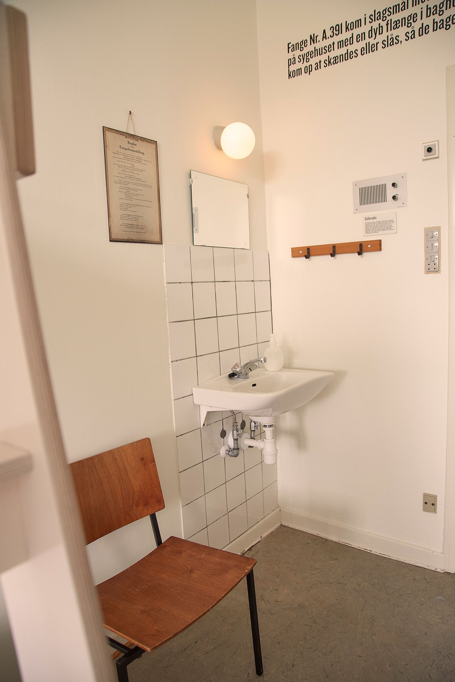 A bathroom with a white sink, a mirror, and a wooden chair. There is a wall-mounted light fixture and a framed certificate on the wall.
