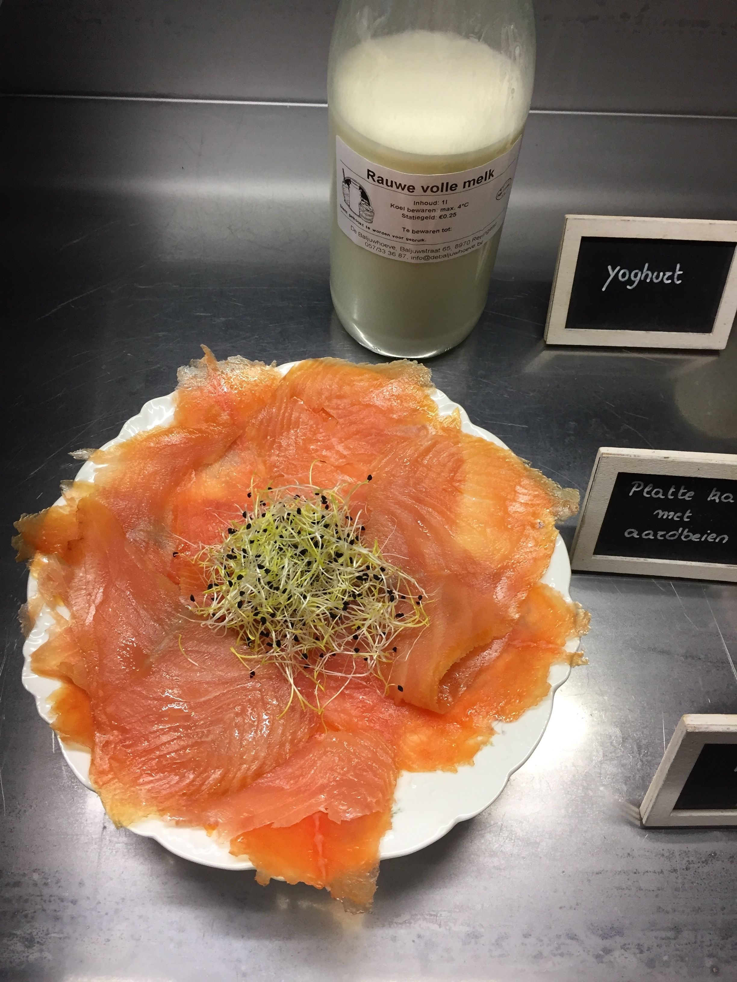 A plate of smoked salmon with sprouts on a white plate, a glass of milk, and signs indicating 'yoghurt' and 'platte met kaasbeien'.