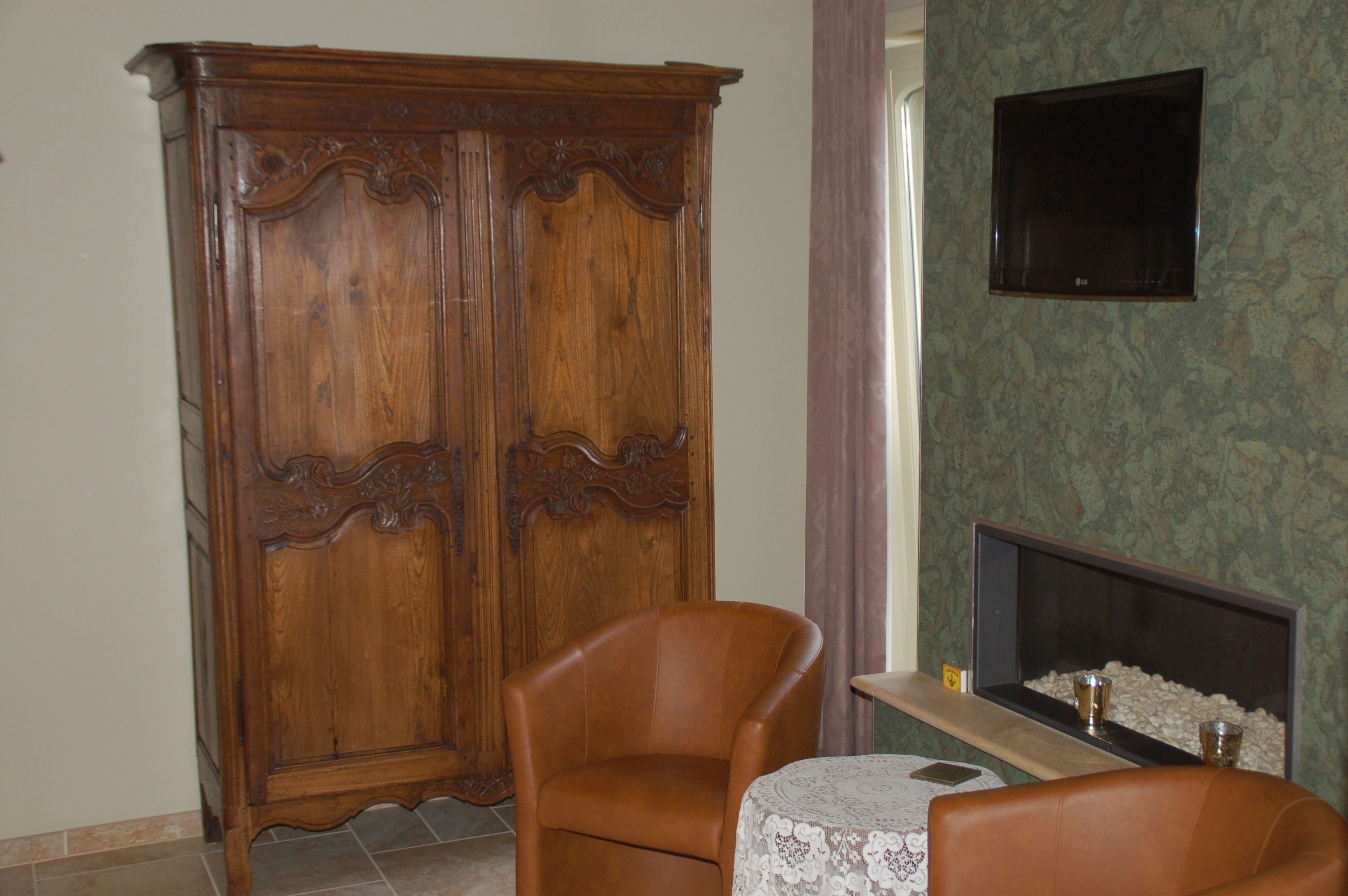 A room with a wooden wardrobe, a leather armchair, a small table with a lace tablecloth, and a flat-screen TV mounted on the wall.