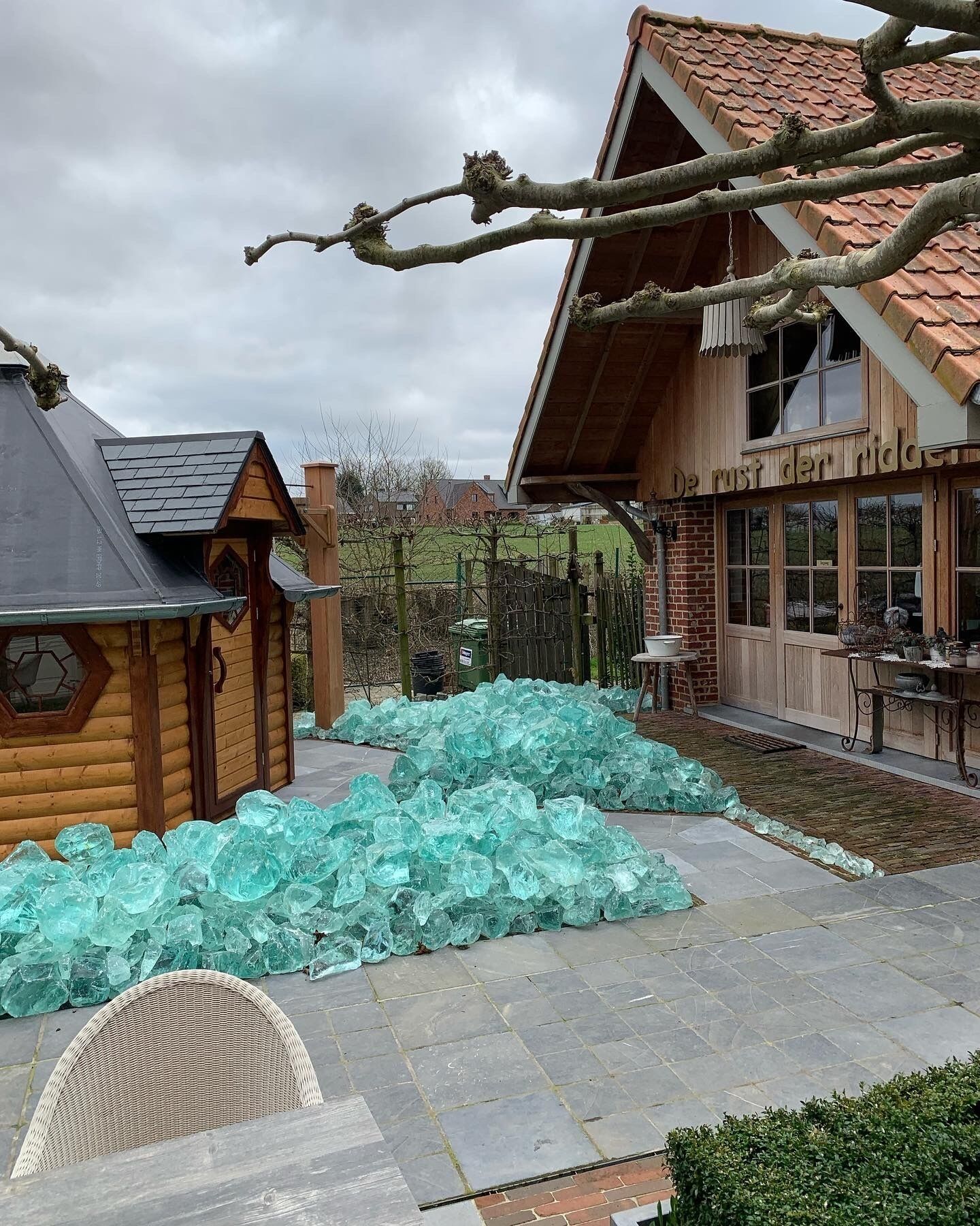 A wooden cabin with a sloped roof, a glass sculpture installation in the yard, and a brick building with large windows.