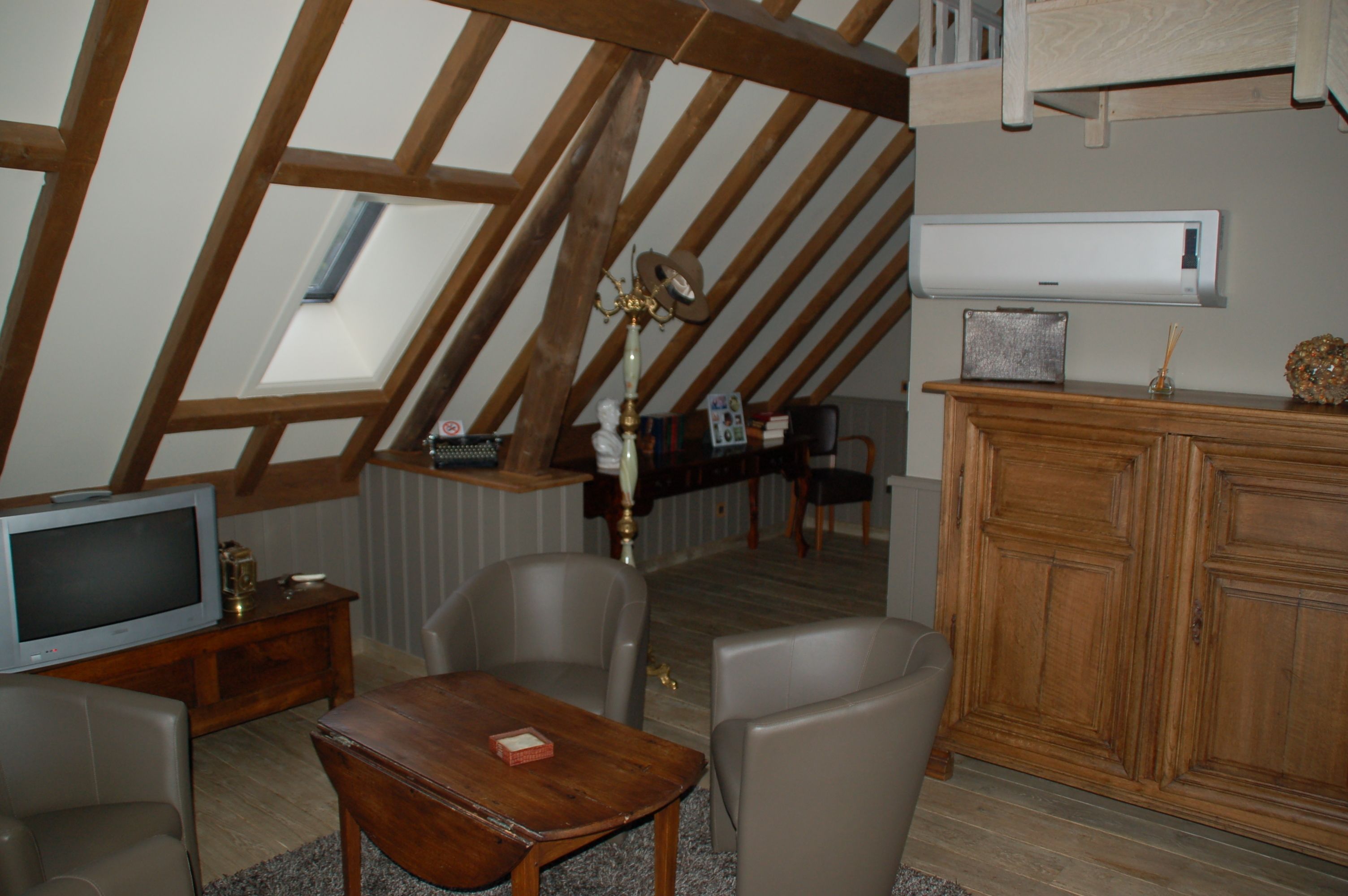 A loft-style room with a sloped ceiling, wooden beams, a television, a small table, two chairs, a cabinet, and a radiator.