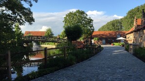 A cobblestone pathway leading to a rustic building with a red-tiled roof, surrounded by greenery and a wooden bridge over a calm water body. - B&B De Rentmeesterhoeve (Poperinge)