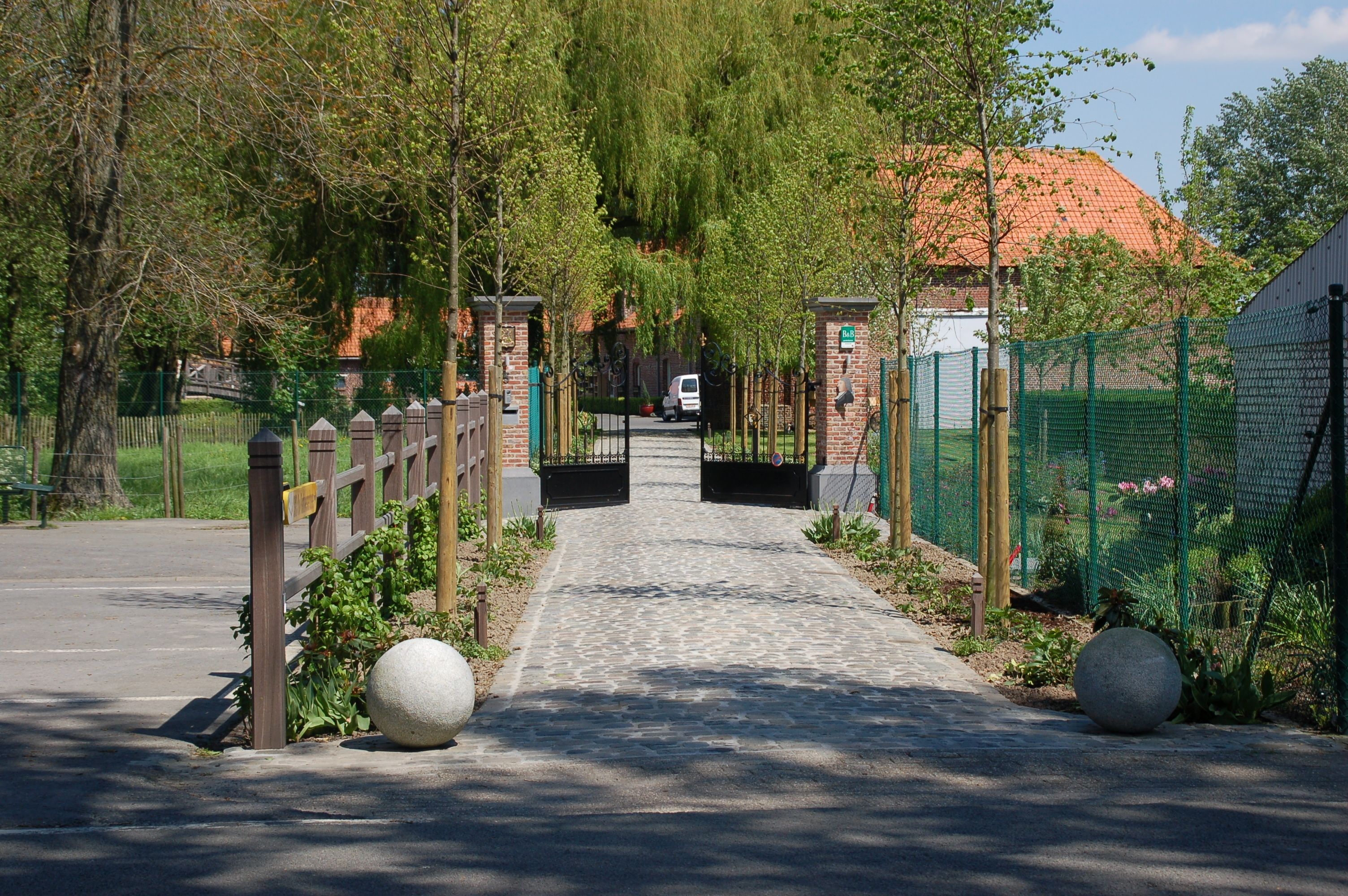 Un chemin pavé mène à une entrée fermée par un portail, adossée à un mur de briques et bordée d’une clôture verte.