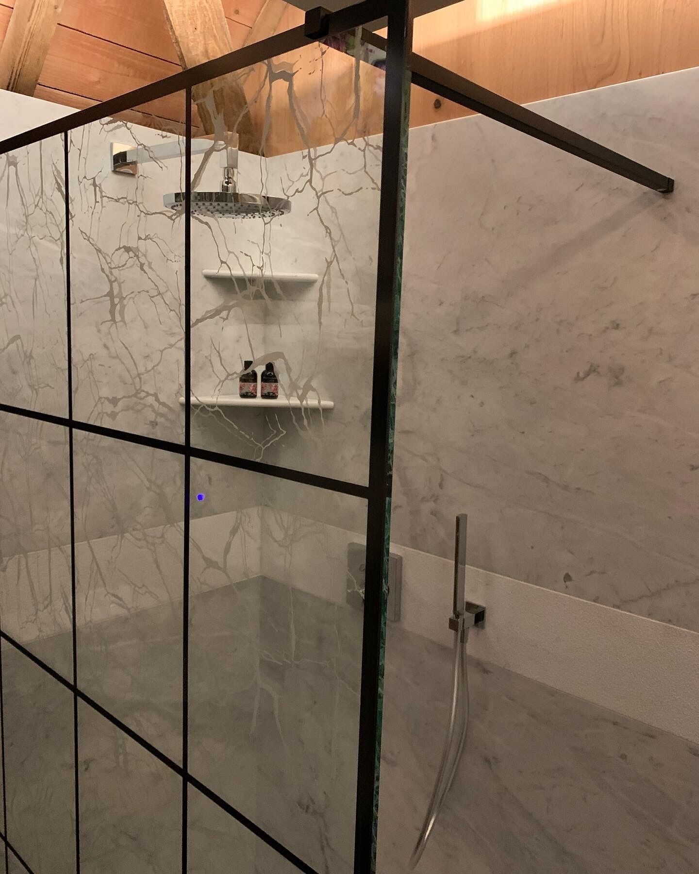 A modern bathroom with a glass shower enclosure, marble walls, and a built-in shelf with toiletries.