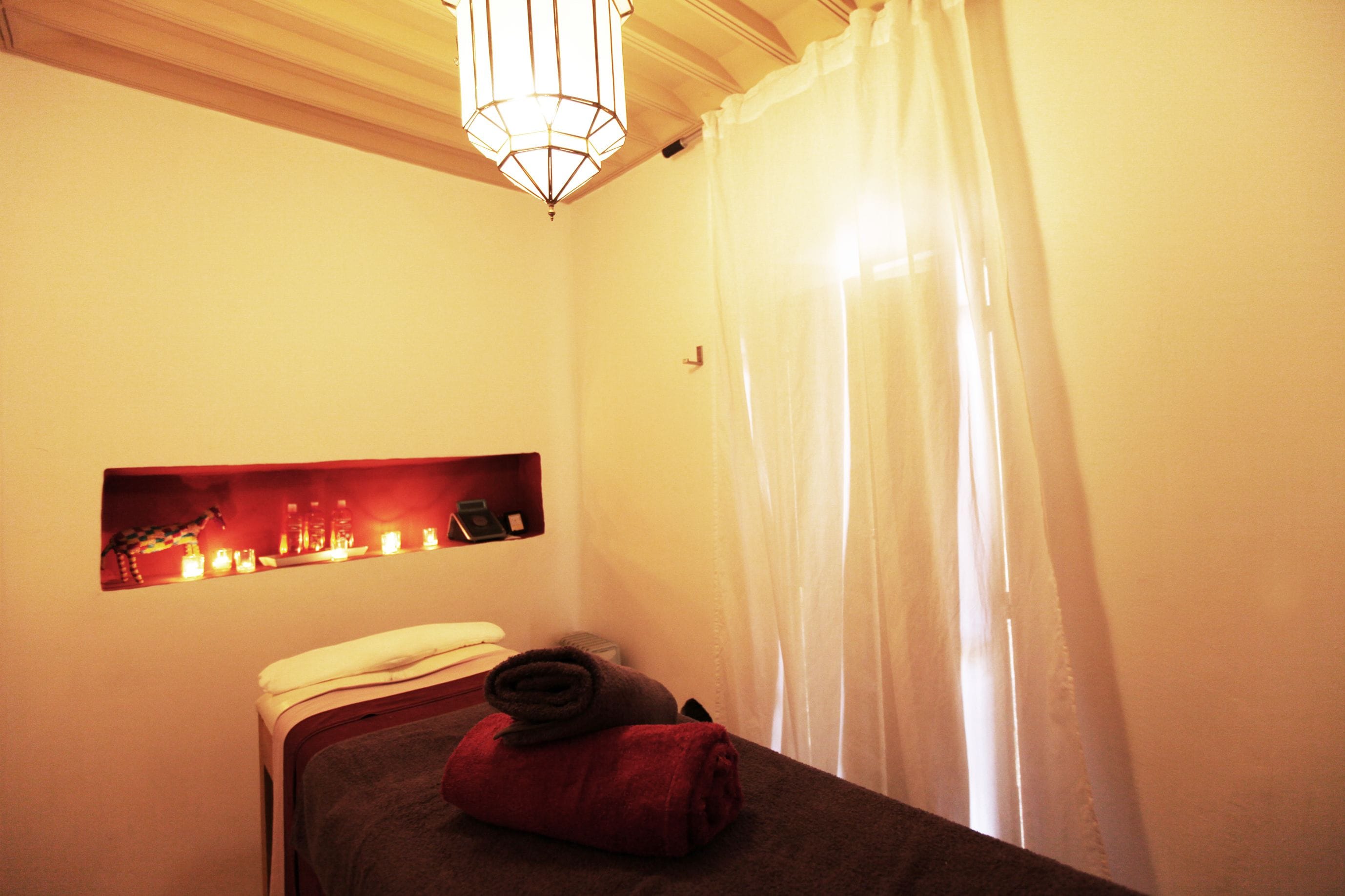 Una habitación de hotel con cama, un pequeño estante con velas y una ventana con cortinas blancas.