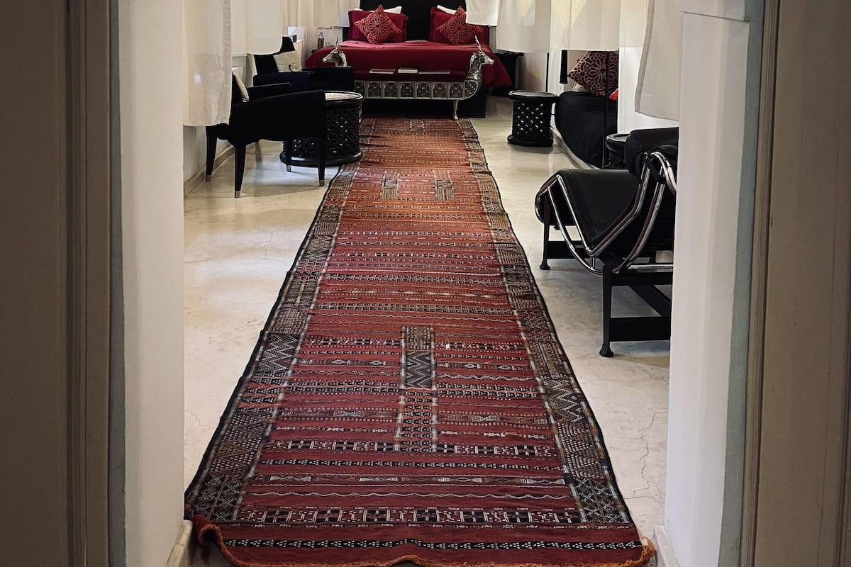 A hallway with a red carpet leading to a bed with a red bedspread and pillows, flanked by black chairs and a small table.