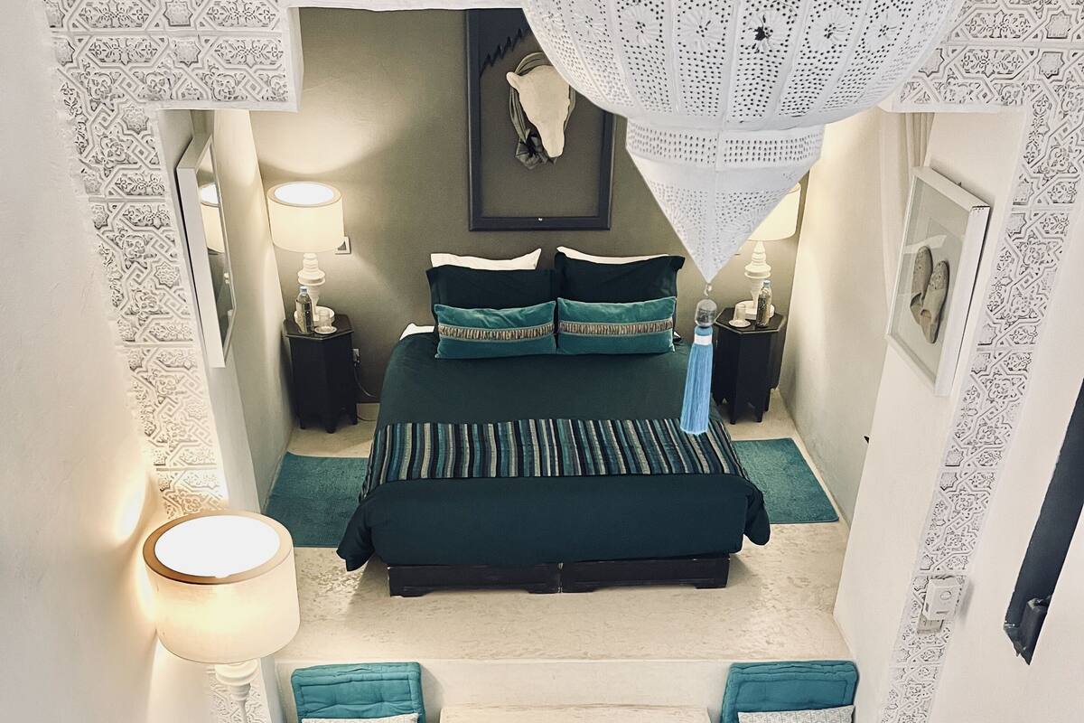 A bedroom with a bed, two nightstands, a lamp, a headboard, a window, and a small seating area with a chair and a round table.