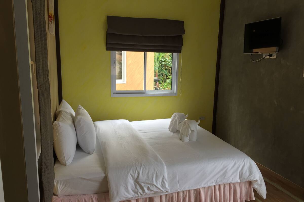 A neatly made bed with white linens and a pink skirted base, a window with a dark valance, and a wall-mounted TV.