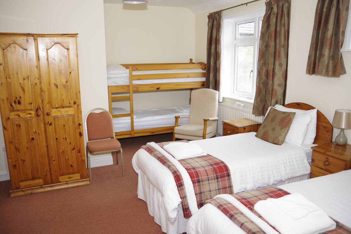 A hotel room with a bunk bed, a single bed, a wooden wardrobe, a chair, a bedside table with a lamp, and a window with curtains.