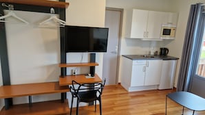 A compact room with a wooden dining table, a black chair, a TV mounted on a wall, a kitchenette with a microwave, and a window with curtains. - Bjugn Hotell (Orland)