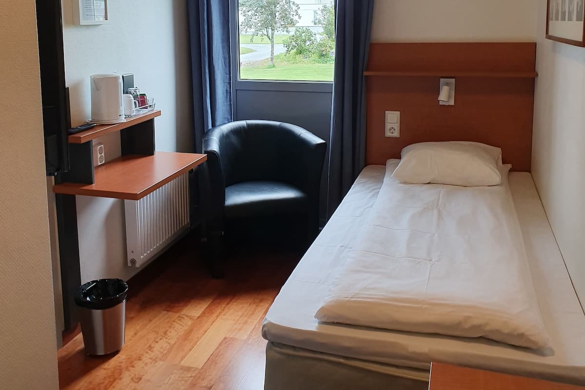 A hotel room with a bed, a chair, a desk with a computer, a radiator, a trash bin, and a window with curtains.
