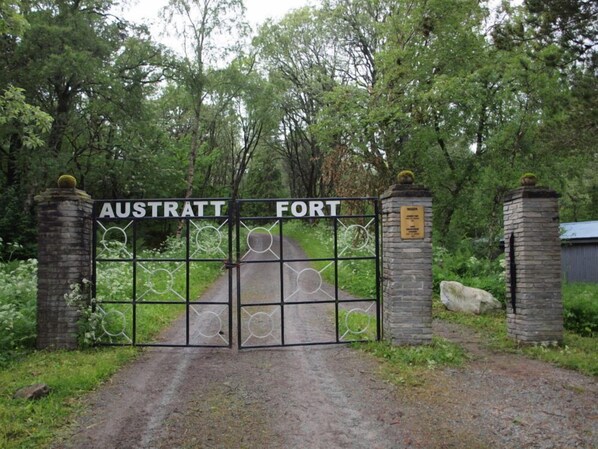 Una puerta con la leyenda 'Austritt Fort', adornada con un diseño metálico decorativo, flanqueada por pilares de piedra y un sendero que atraviesa un área boscosa.