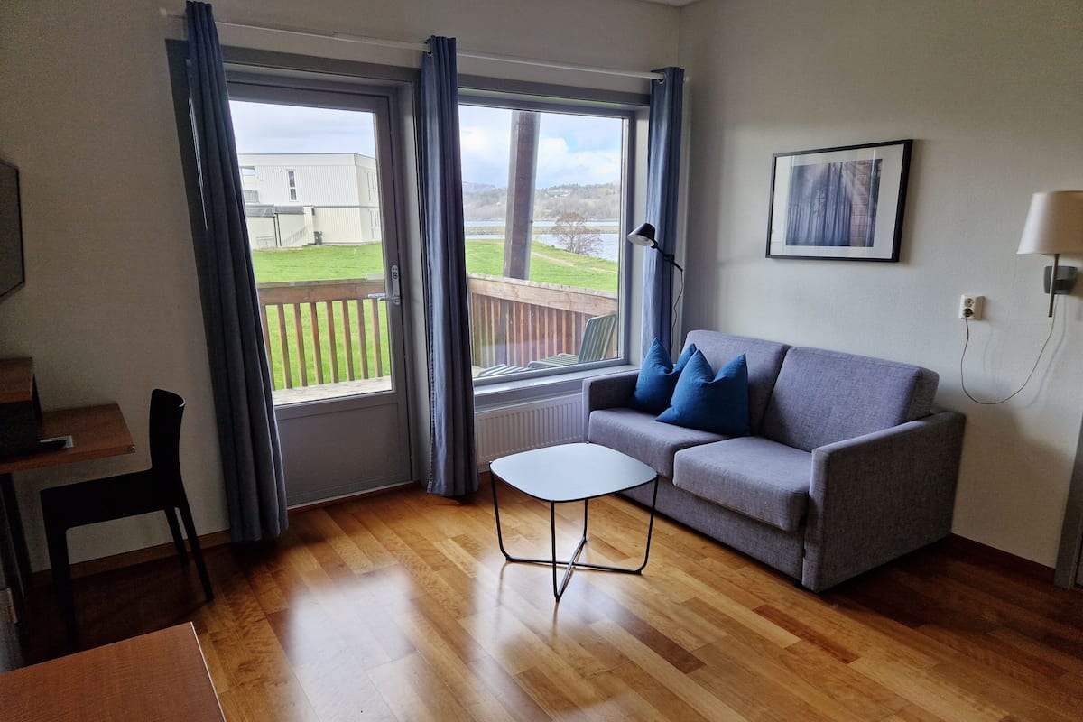 A room with a grey sofa, a small white table, wooden flooring, a window with curtains, a framed picture on the wall, and a view of a building and greenery outside.