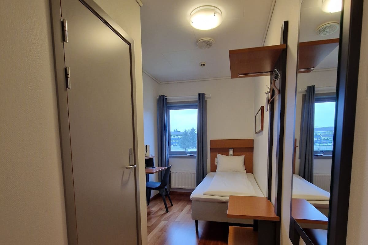 A hotel room with a bed, a desk, a chair, a mirror, and a window with a view of the outside.