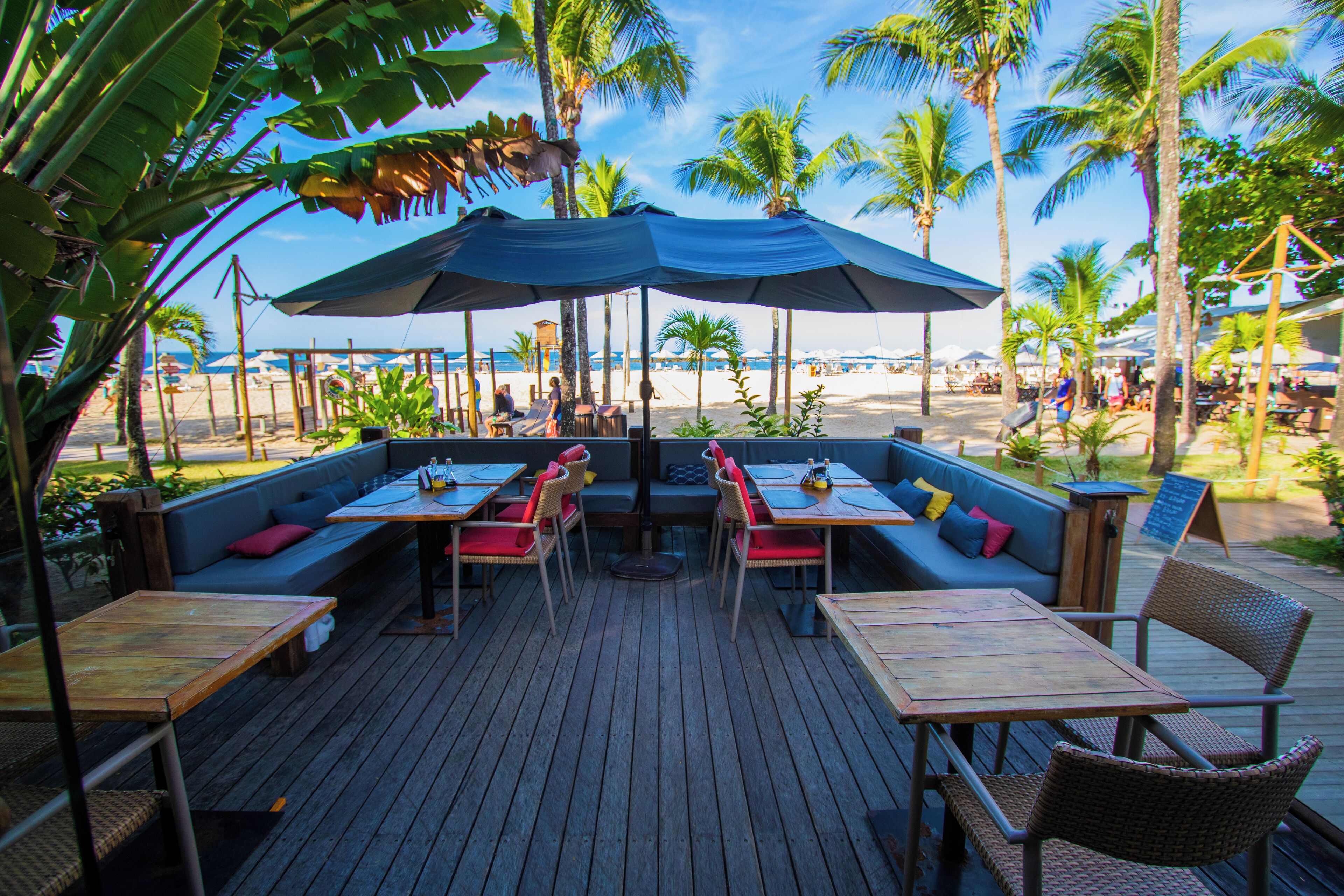 An outdoor dining area with wooden tables and chairs, a blue canopy, and a view of the beach and palm trees.