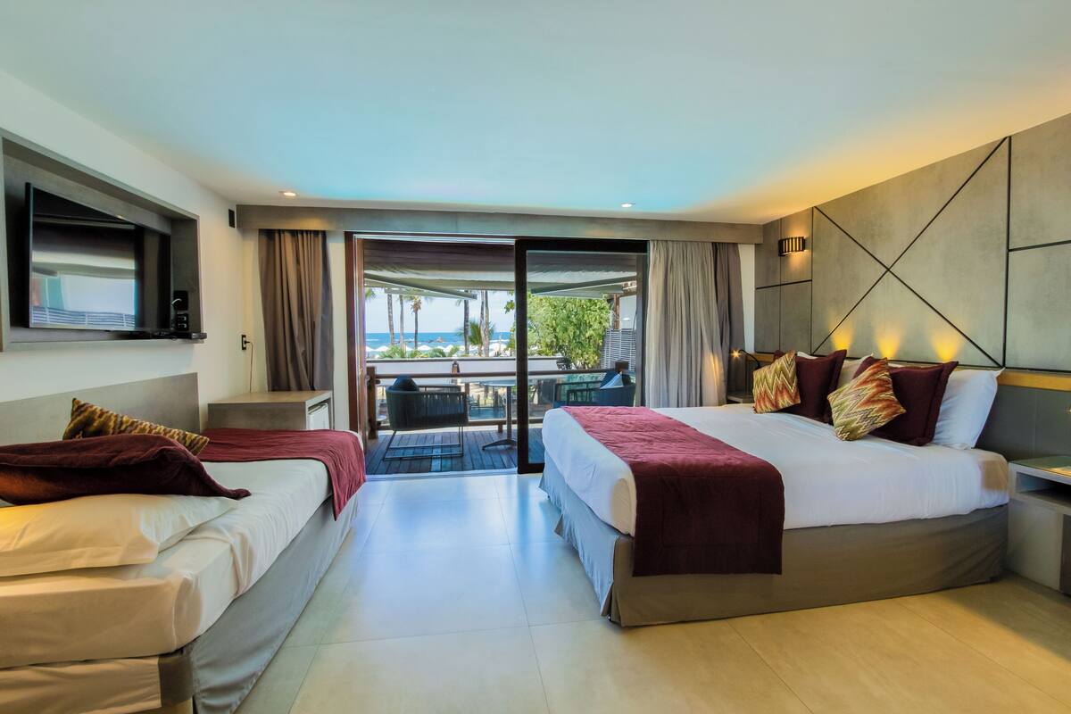 A modern hotel room with two beds, a balcony with a view of the beach, and a flat-screen TV.