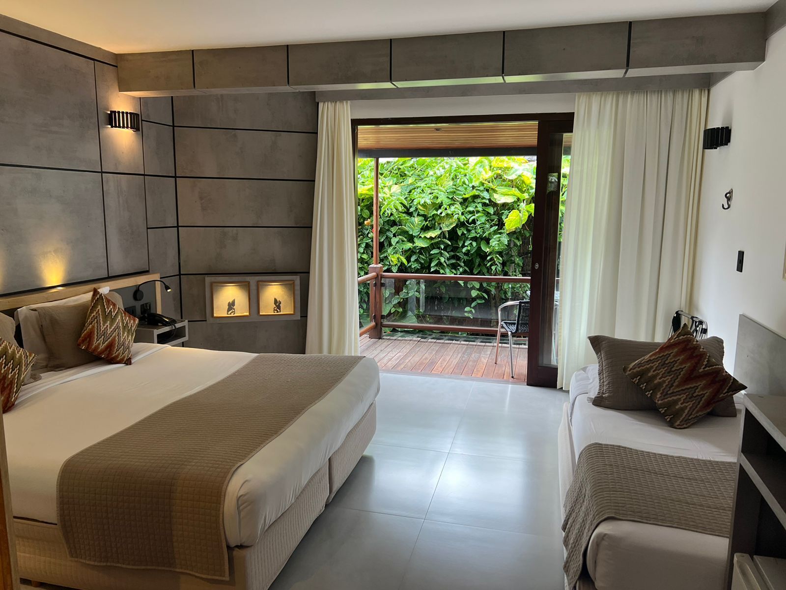 Una habitación de hotel moderna con una cama grande, una cama más pequeña, un balcón con vistas a la vegetación y una zona de estar.