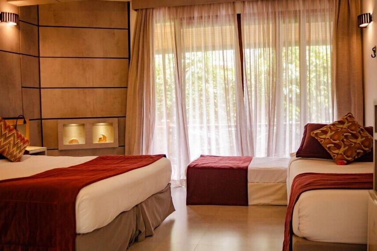A hotel room with two beds, a large window with sheer curtains, and wall-mounted lamps.