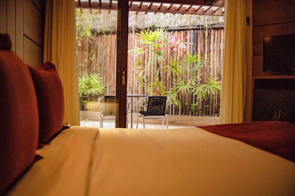 A room with a bed, a chair, and a view of a bamboo fence and plants outside.