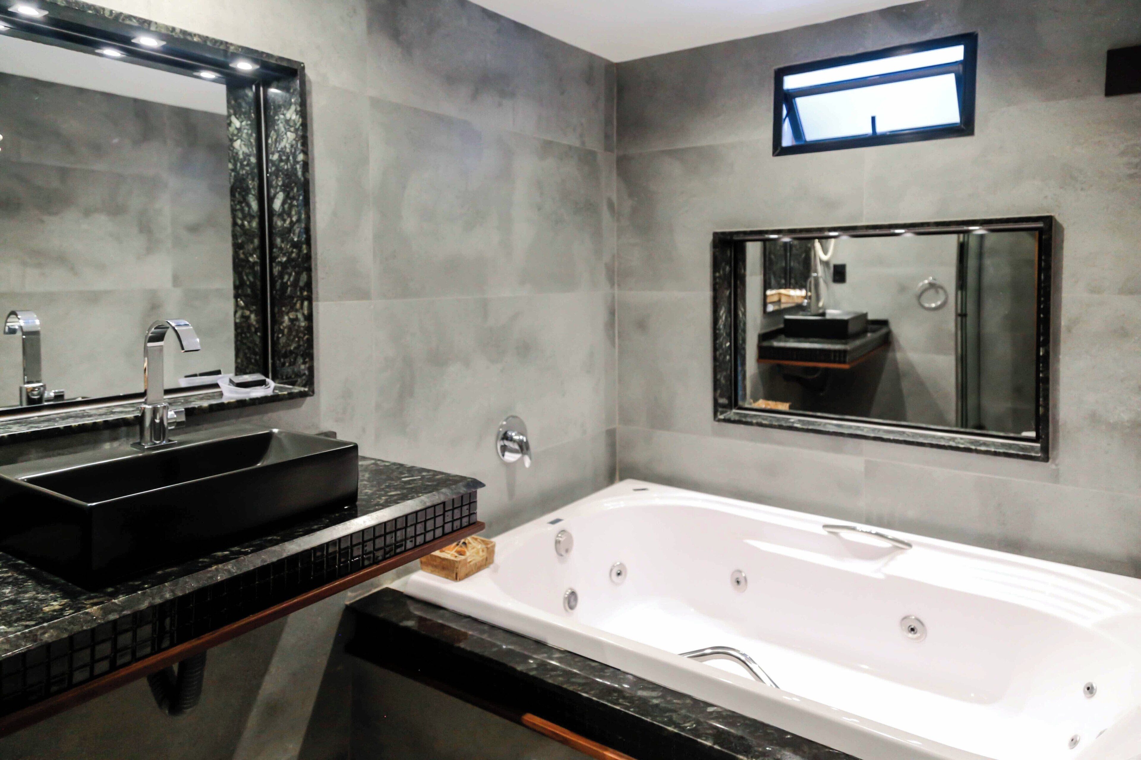 A modern bathroom with a large mirror, a black sink, a white bathtub, and a small window.