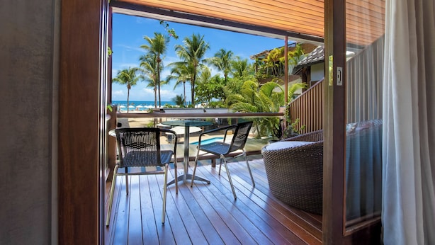 A wooden deck with a glass balustrade overlooking a pool and palm trees, featuring outdoor seating and a view of the ocean. - Pousada da Torre (Cairu)