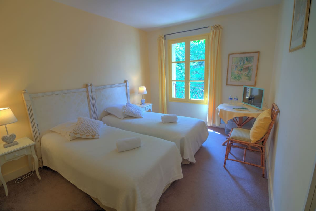 A room with two beds, a small table with a chair, a window with curtains, and a painting on the wall.