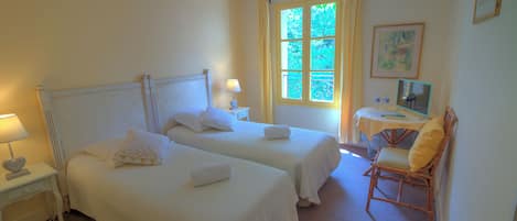 A room with two beds, a small table with a chair, a window with curtains, and a painting on the wall.