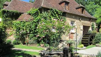 Ein Haus aus Stein mit traditionellem Satteldach, Wasserrad und einem Garten mit blühenden Pflanzen.