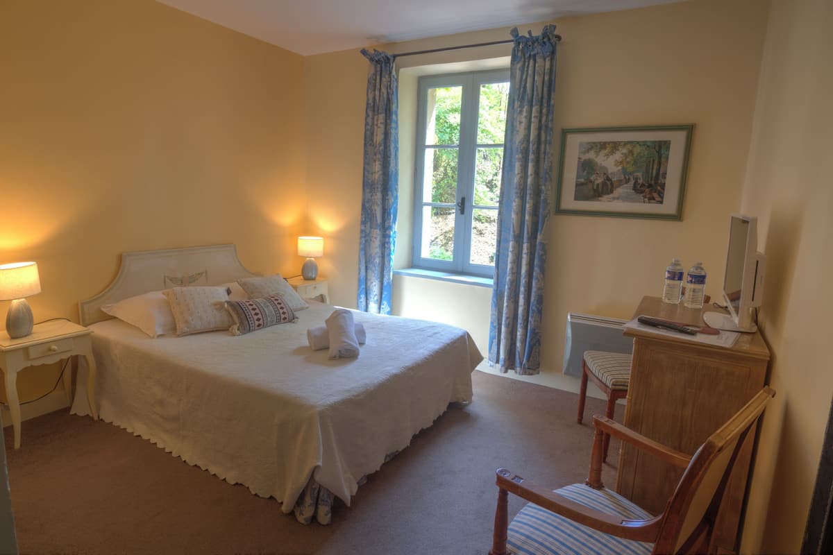 A bedroom with a bed, bedside lamps, a chair, a desk, a window with curtains, and a painting on the wall.