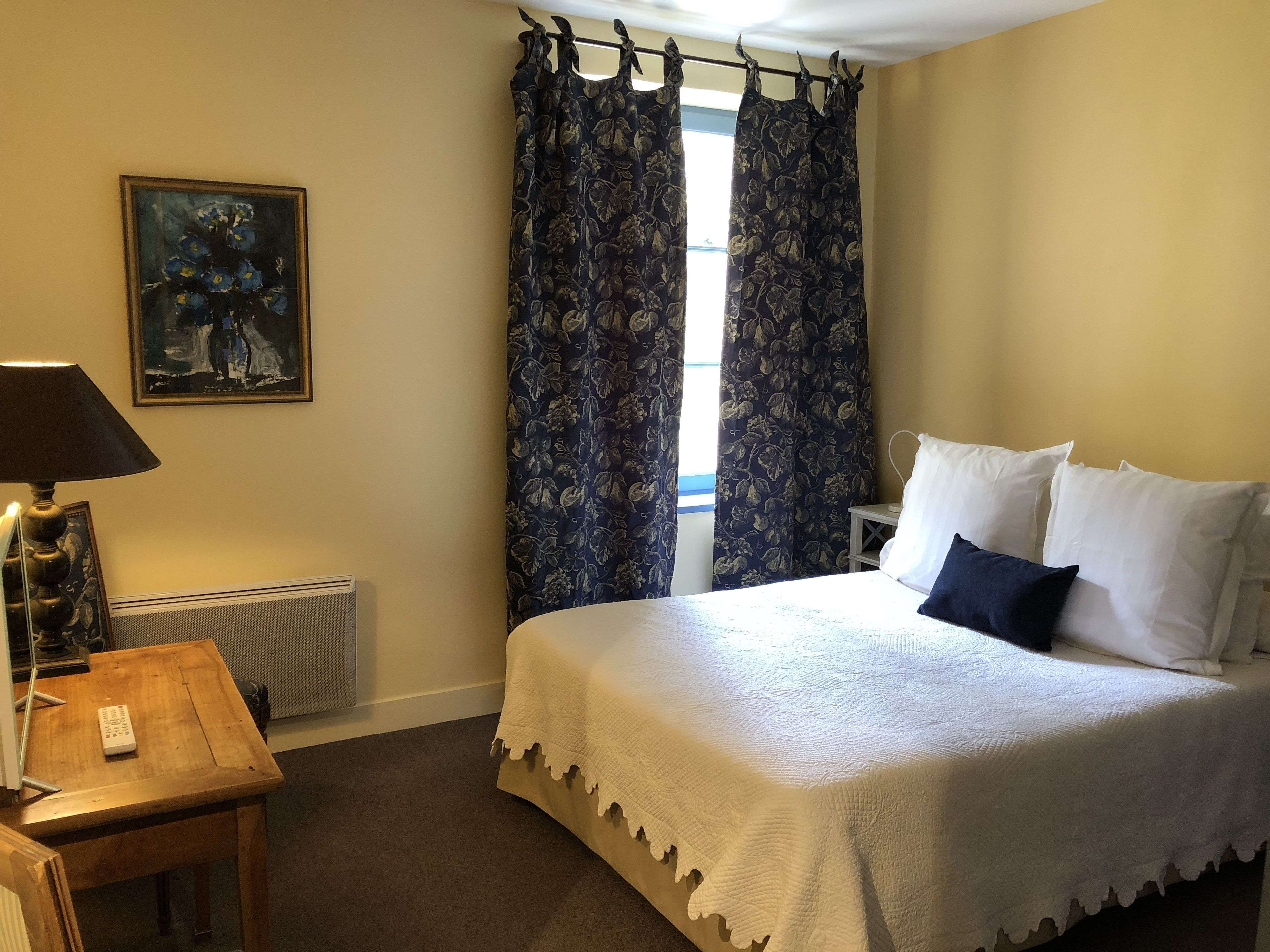 A hotel room with a bed, a desk, a lamp, a painting, and a window with curtains.