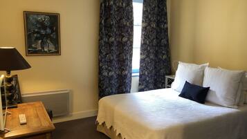 A hotel room with a bed, a desk, a lamp, a painting, and a window with curtains.