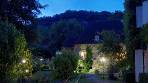 Ein Steinhaus mit einem geneigten Dach, umgeben von einem gepflegten Garten mit Bäumen und Sträuchern. Im Vordergrund stehen zwei Liegestühle.