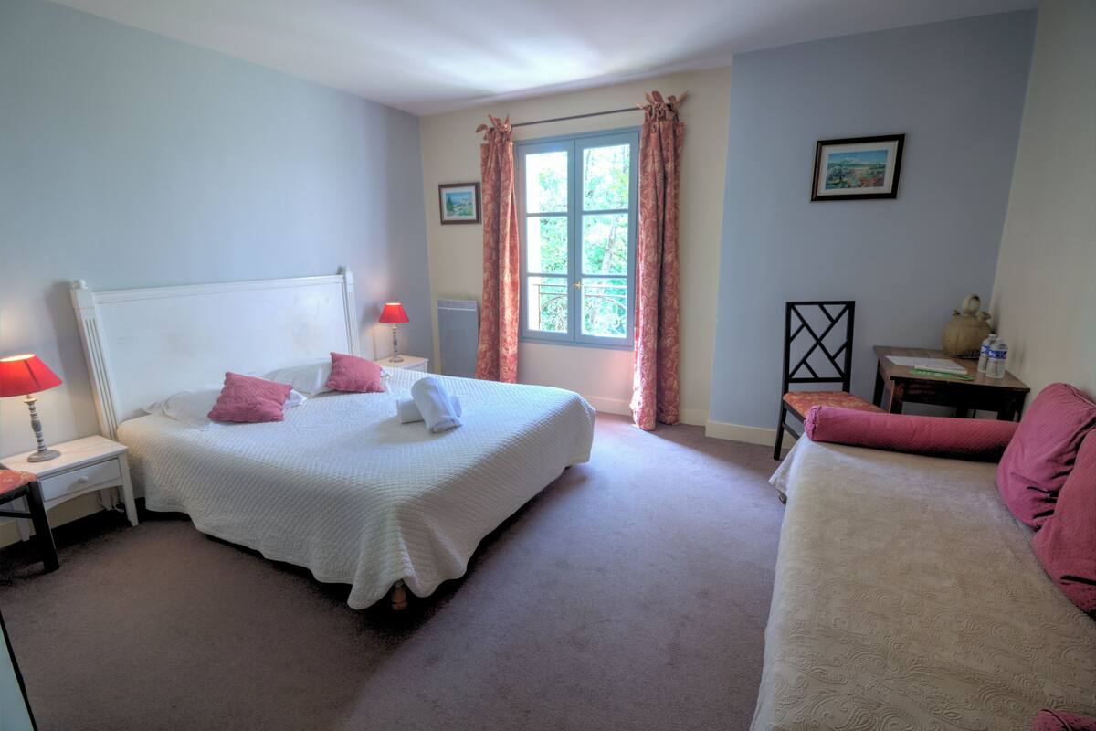 A bedroom with a bed, bedside tables, a window with curtains, a chair, and a small table with a lamp.