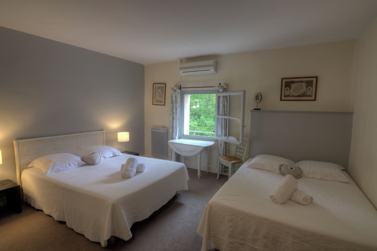A hotel room with two beds, a small table, and a window with a view of greenery.