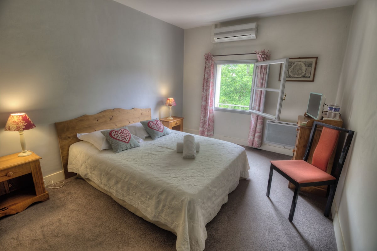 A bedroom with a bed, bedside lamps, a chair, a desk with a computer, and a window with curtains.
